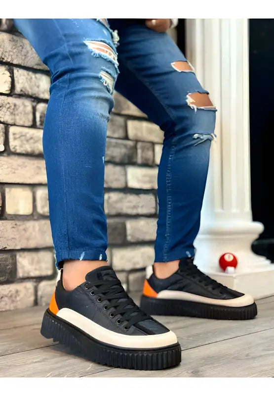 Black platform sneakers with orange accents and white soles worn with ripped blue jeans.