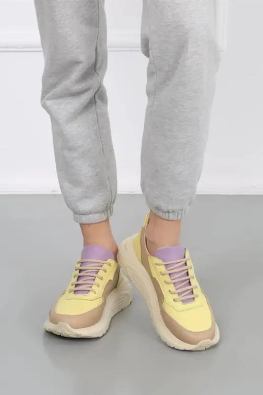 Yellow and beige chunky sneakers with purple laces worn with light gray pants.