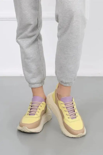 Yellow and beige chunky sneakers with purple laces worn with light gray pants.