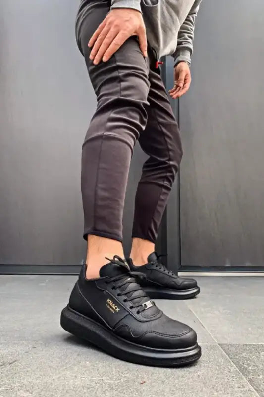 Black leather sneakers with thick soles and gold accents worn with gray pants.