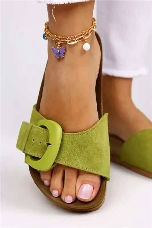 Lime green suede sandal with a prominent buckle and an ankle chain bracelet with butterfly charm.