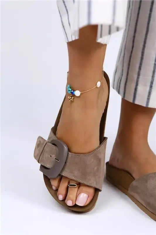 Taupe suede sandal with a prominent square buckle and an ankle chain accessory.