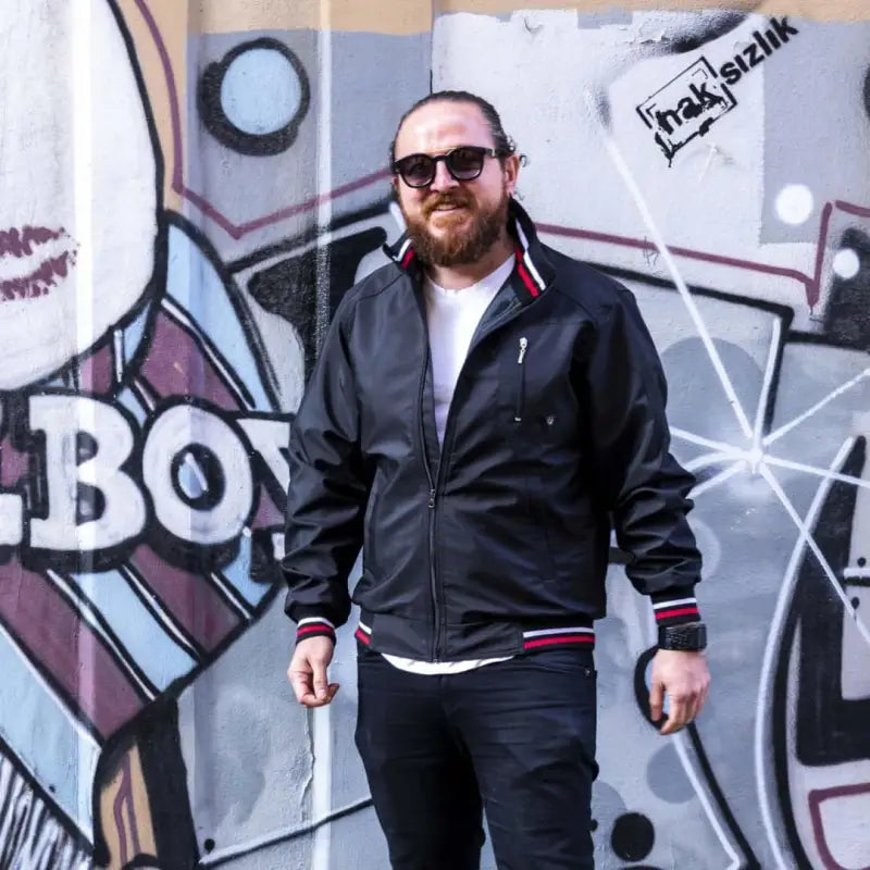 A bearded person wearing sunglasses, a black track jacket, and dark pants standing in front of graffiti.